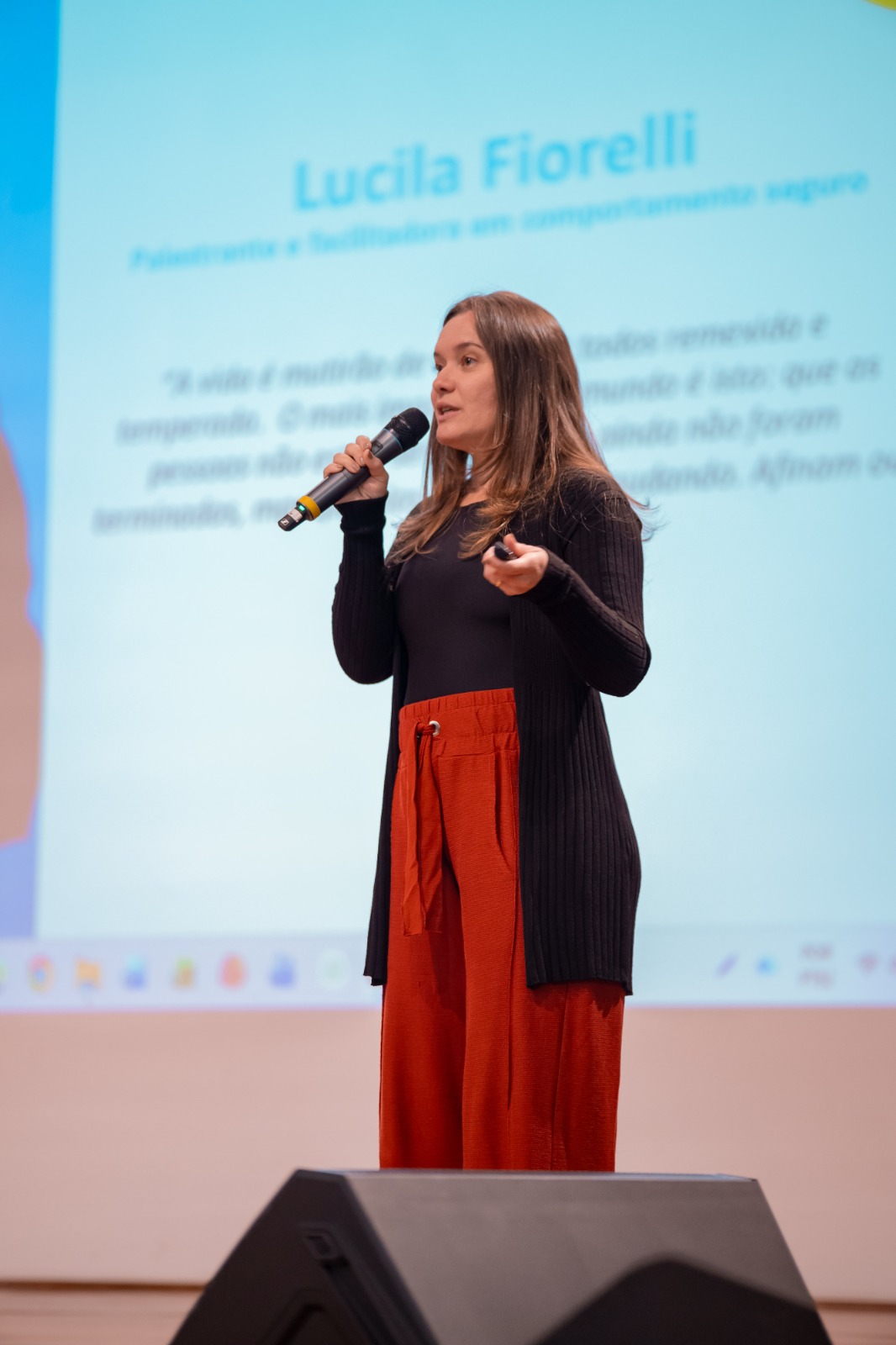 Lucila Fiorelli em pé no palco, com microfone na mão, cardigã preto e pantalona terracota, diante de um telão azul com o nome dela